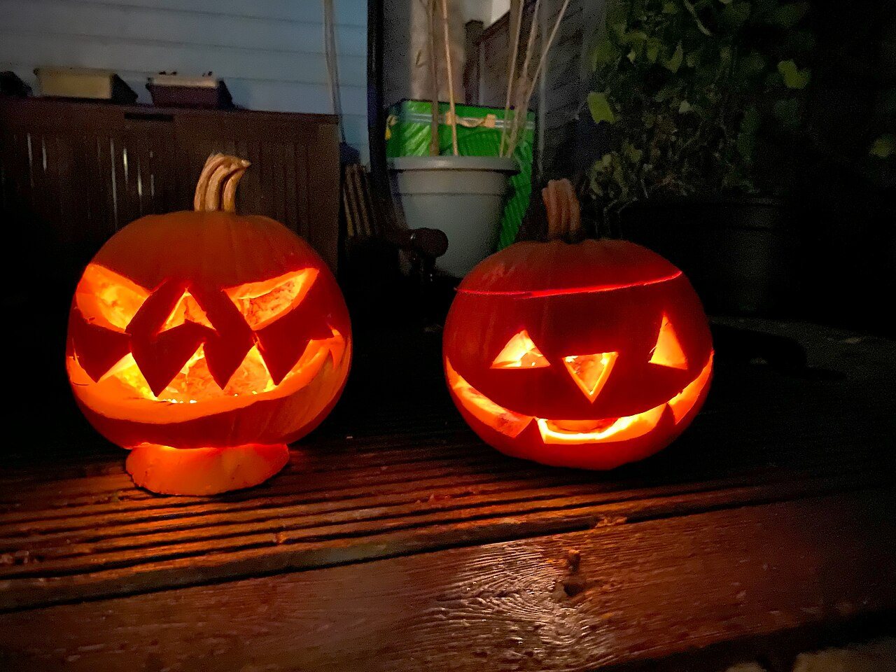 Two carved pumpkin jack-o'-lanterns glowing orange on a wooden deck at night. Photo: Lirazelf, CC BY-SA 4.0, via Wikimedia Commons
