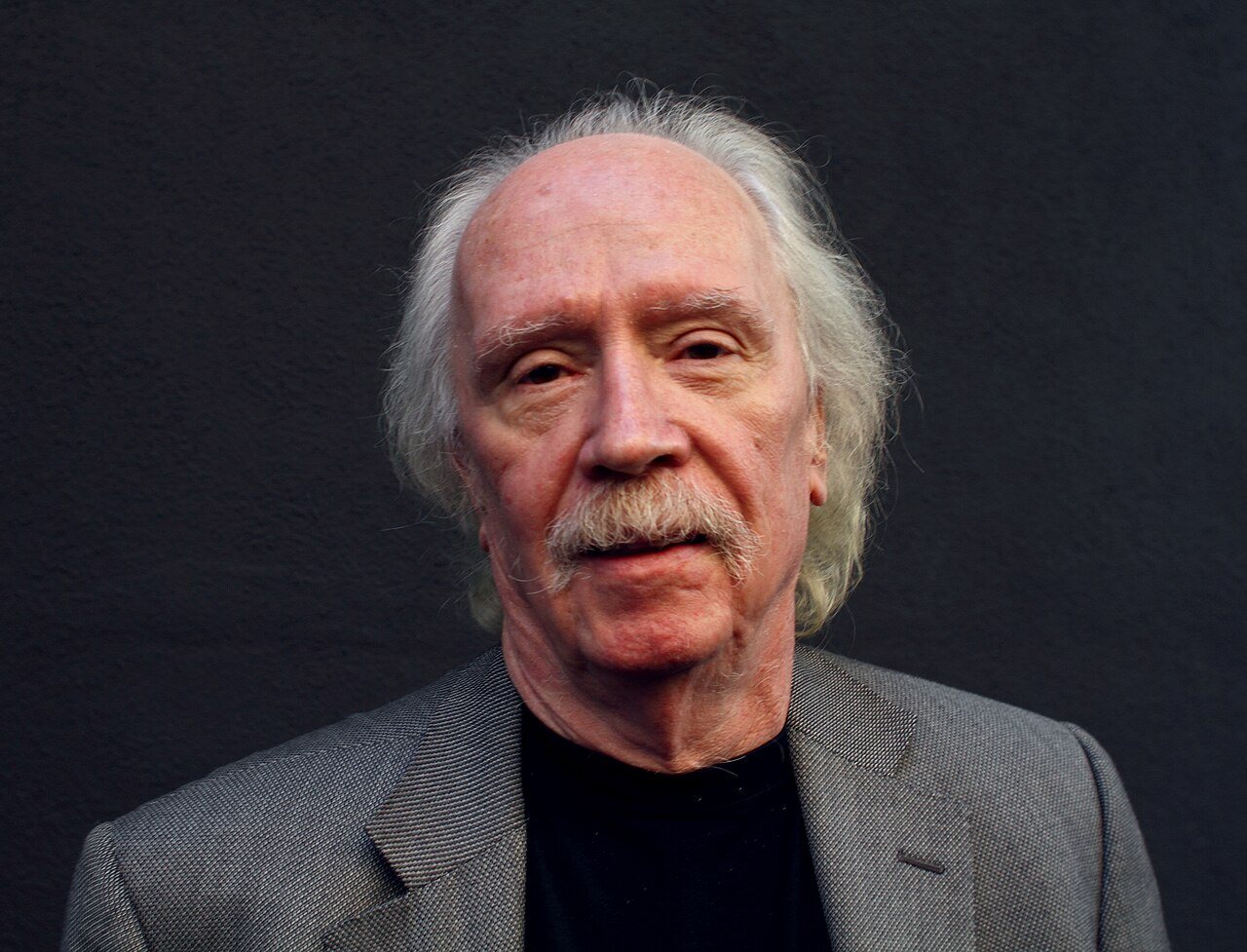 Director John Carpenter, with long white hair and a gray moustache, in a gray jacket against a dark wall in 2010